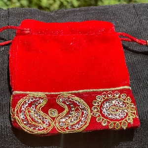 May include: A red velvet drawstring pouch with a gold and silver embroidered border. The pouch is decorated with a paisley pattern.