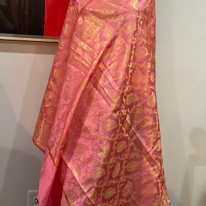 May include: A pink and gold patterned silk scarf with a floral design. The scarf has a fringed edge and is draped over a mannequin.