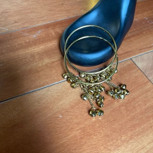 May include: A pair of gold hoop earrings with dangling charms. The charms are small, round, and have a bell-like shape.