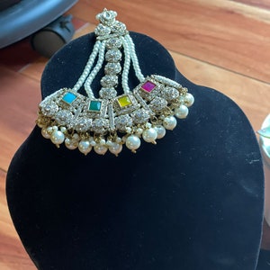 May include: A gold and white bridal headpiece with a pearl and crystal design. The headpiece features a cluster of colorful stones in the center.