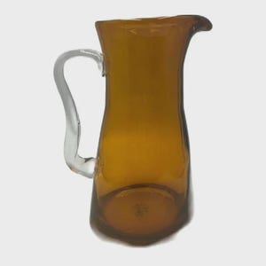 Op de afbeelding: Amberkleurige glazen kan met een helderglazen handvat. De kan heeft een uitlopende bovenkant en een afgeronde basis. Het handvat is gebogen en aan de zijkant bevestigd. Deze vintage kan is geschikt voor het serveren van dranken.