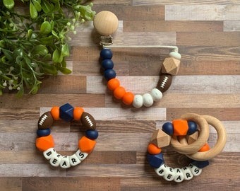 May include: Three silicone teething toys in orange, blue, and white with the word "BEARS" spelled out in white beads. The toys are shaped like a ring, a football, and a rattle with wooden rings.