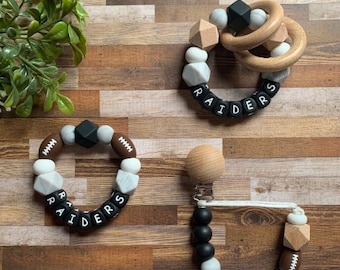 May include: Three silicone teething toys with the word "RAIDERS" spelled out in black letters. The toys are made with white and brown silicone beads and feature a football design. One toy is a ring with a wooden ring, another is a bracelet, and the third is a clip-on teether.
