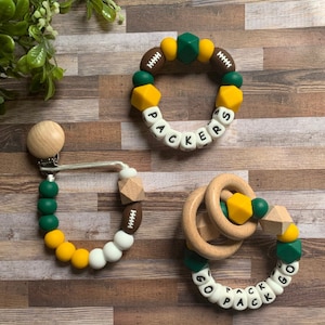 May include: Three silicone teethers in green, yellow, and white with brown accents. One teether is a bracelet with the word "PACKERS" spelled out. Another teether is a ring with the words "GO PACK GO" spelled out. The third teether is a clip-on teether with a football design.