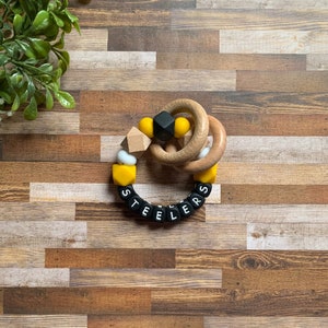 May include: A black and yellow silicone teething ring with the word "STEELERS" spelled out in black beads. The ring is attached to a wooden ring.