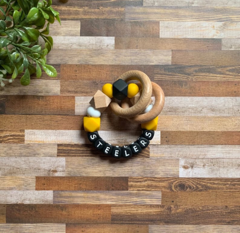 May include: A black and yellow silicone teething ring with the word "STEELERS" spelled out in black beads. The ring is attached to a wooden ring.
