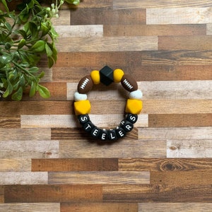 May include: A black, yellow, and brown silicone teething bracelet with the word "STEELERS" spelled out in black letters. The bracelet features football-shaped beads and a black square bead.