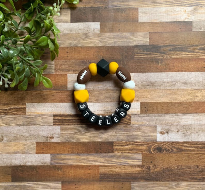 May include: A black, yellow, and brown silicone teething bracelet with the word "STEELERS" spelled out in black letters. The bracelet features football-shaped beads and a black square bead.