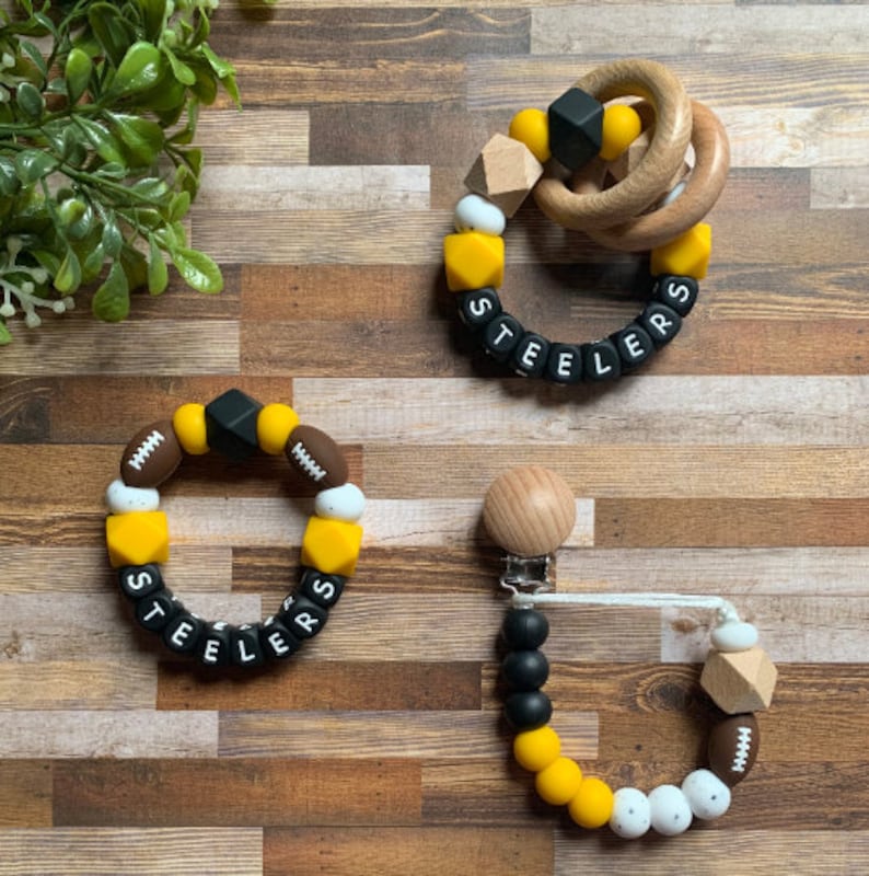 May include: Three silicone teething toys with the word "Steelers" spelled out in black letters. The toys are in the colors black, yellow, white, and brown. One toy is a ring with a wooden ring attached. Another toy is a bracelet with footballs and a diamond shape. The third toy is a clip-on teether with a wooden bead.