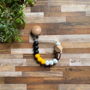 May include: A yellow, black, and white silicone baby teether with a wooden clip and a football charm. The teether is strung on a white cord.