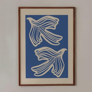 May include: A framed art print featuring two abstract, cream-colored floral shapes on a blue background. The print hangs above a table set with a blue mug, small orange plate, a basket of bread rolls, and two lemons. The table is covered with a white tablecloth with black polka dots.