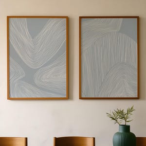 Peut inclure: Deux impressions encadrées d'art abstrait avec des motifs de lignes blanches sur fond bleu clair. Les cadres sont de couleur marron chaud. Un vase vert foncé avec une plante et une table en bois avec une assiette vert foncé sont également visibles.