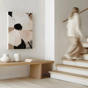 May include: Abstract floral canvas art in neutral tones of white, black, and beige hangs on a white wall. A wooden bench holds two white vases and two books. A person walks down a wooden staircase.