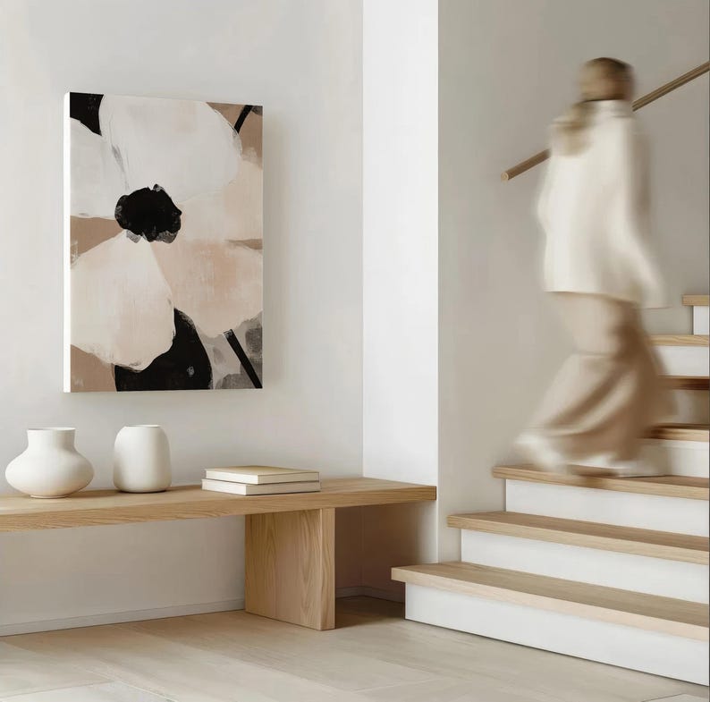 May include: Abstract floral canvas art in neutral tones of white, black, and beige hangs on a white wall. A wooden bench holds two white vases and two books. A person walks down a wooden staircase.