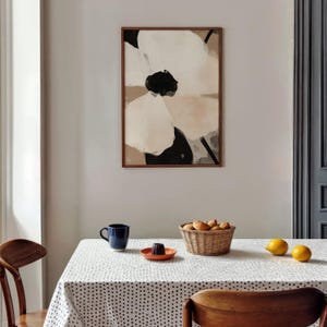 May include: A framed abstract floral painting in neutral tones hangs above a table set with a blue mug, orange plate, and a wicker basket of bread rolls. Two lemons sit on the dotted tablecloth. The painting features white, black, and beige brushstrokes.
