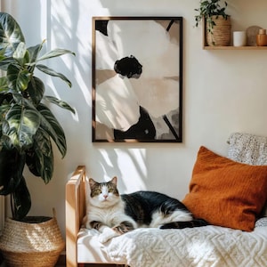 May include: A framed abstract floral art print in neutral tones hangs above a wooden bench. An orange pillow and a white blanket are on the bench. A tabby cat is resting on the bench. A large potted plant is to the left.