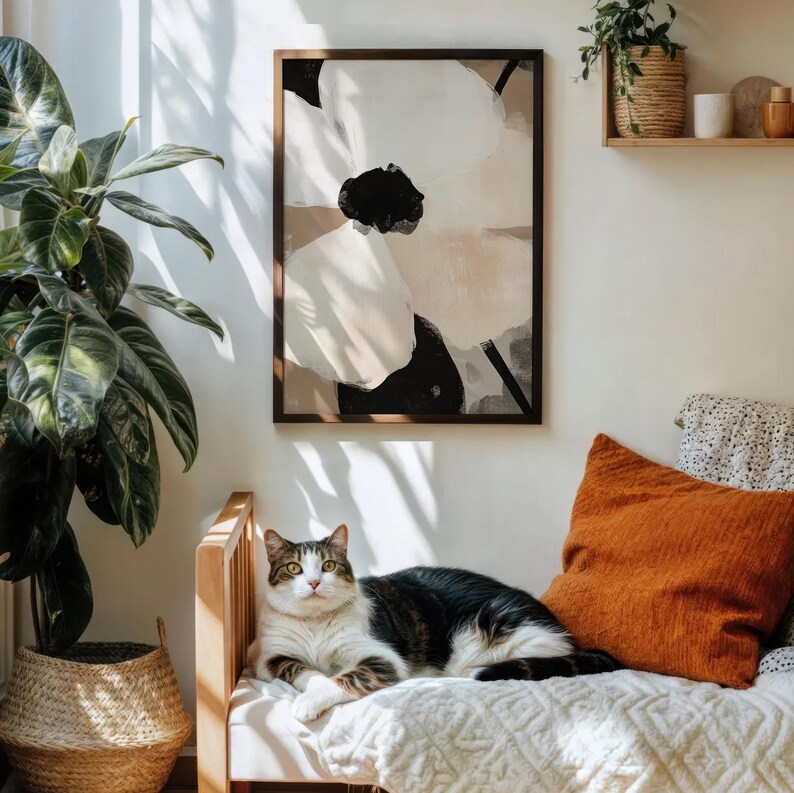 May include: A framed abstract floral art print in neutral tones hangs above a wooden bench. An orange pillow and a white blanket are on the bench. A tabby cat is resting on the bench. A large potted plant is to the left.