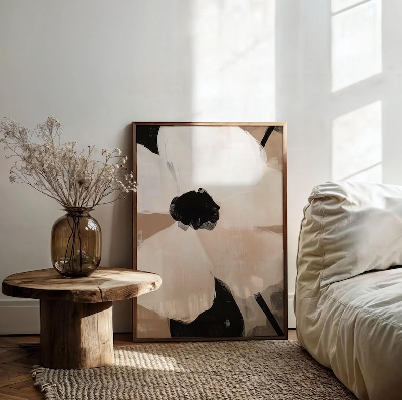 May include: A framed abstract painting of a large white flower with black and brown accents on a beige background. The painting is hanging on a white wall next to a wooden table with a vase of dried flowers.