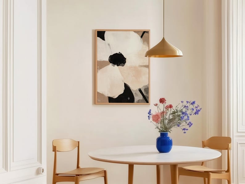 May include: A framed abstract floral art print with beige, black, and white colors hangs above a round white table. A blue vase holds a bouquet of flowers. Two wooden chairs are positioned near the table, and a gold pendant light hangs from the ceiling.