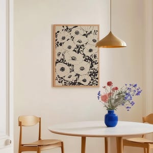 K&ouml;nnte beinhalten: Ein gerahmtes Kunstwerk mit einem schwarz-wei&szlig;en Blumendruck h&auml;ngt &uuml;ber einem runden wei&szlig;en Tisch mit zwei Holzst&uuml;hlen. Eine blaue Vase mit rosa und violetten Blumen steht auf dem Tisch. Eine goldene Pendelleuchte h&auml;ngt &uuml;ber dem Tisch.