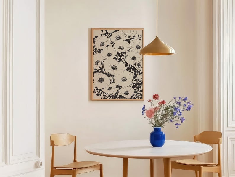 K&ouml;nnte beinhalten: Ein gerahmtes Kunstwerk mit einem schwarz-wei&szlig;en Blumendruck h&auml;ngt &uuml;ber einem runden wei&szlig;en Tisch mit zwei Holzst&uuml;hlen. Eine blaue Vase mit rosa und violetten Blumen steht auf dem Tisch. Eine goldene Pendelleuchte h&auml;ngt &uuml;ber dem Tisch.