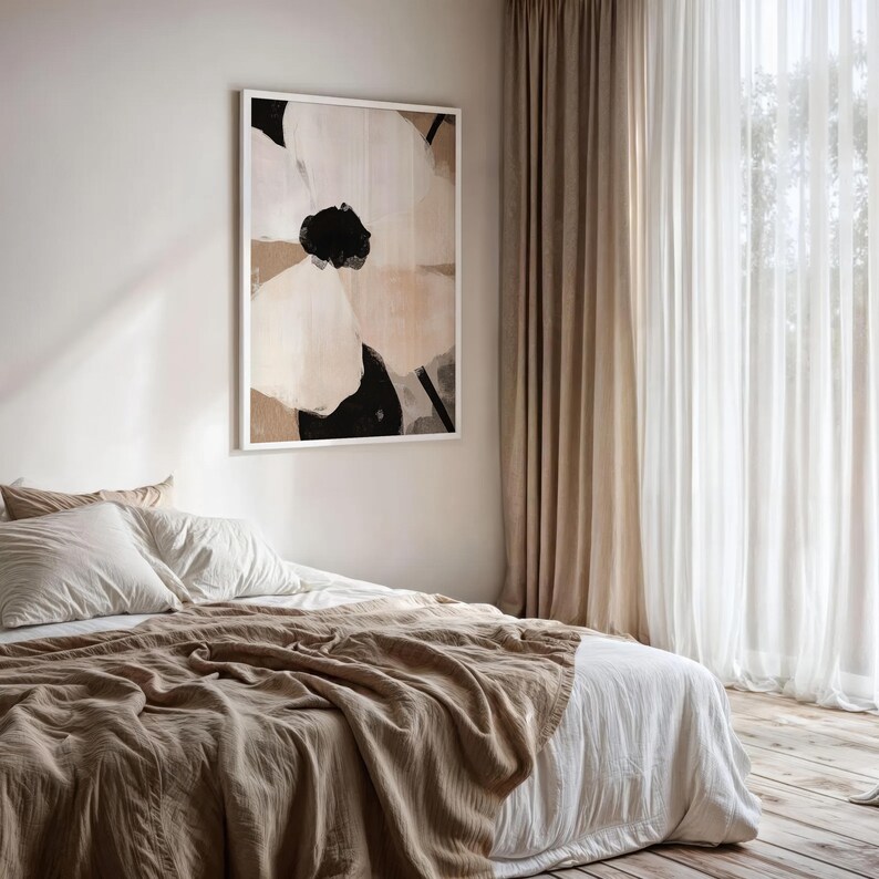 May include: Abstract floral art print in a white frame. The artwork features large, overlapping shapes in black, beige, and white. The print hangs above a bed with white linens and a tan throw blanket. Curtains frame a window.