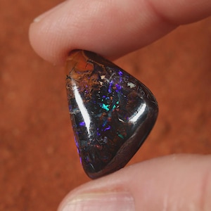 Könnte beinhalten: Ein polierter, dreieckiger schwarzer Opal-Edelstein mit lebendigen Blitzen in Blau, Grün und Lila. Der Opal wird zwischen zwei Fingern gehalten und zeigt sein schillerndes Farbspiel vor einem warmen, braunen Hintergrund. Der Stein hat eine glänzende, reflektierende Oberfläche.