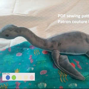 Peut inclure: Un jouet en peluche gris en forme de plésiosaure, un reptile marin préhistorique au long cou et aux nageoires. Le jouet est posé sur un tissu bleu à motif marin. Le texte "PDF sewing pattern Patron couture PDF" est visible dans le coin supérieur droit de l'image. Une échelle de difficulté est représentée sous le jouet avec quatre cercles de couleur : bleu, vert, jaune et rose.