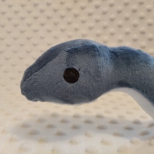 May include: Close-up of a blue plush snake with a black eye. The snake is lying on a white textured surface.