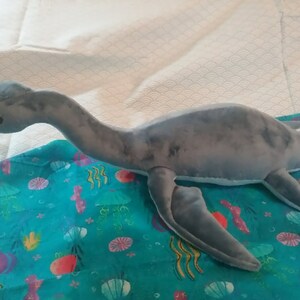 May include: A gray plush toy shaped like a plesiosaur, a long-necked marine reptile, with a long tail and flippers. The toy is lying on a blue fabric with a sea-life pattern.