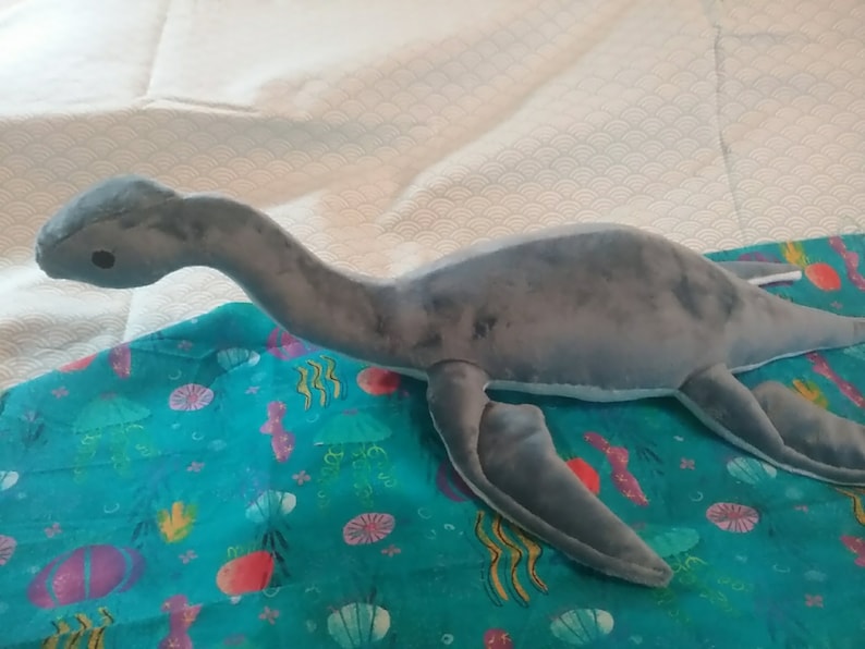 May include: A gray plush toy shaped like a plesiosaur, a long-necked marine reptile, with a long tail and flippers. The toy is lying on a blue fabric with a sea-life pattern.