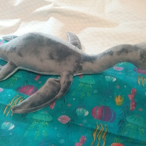 May include: A gray plush toy shaped like a plesiosaur, a long-necked marine reptile, lying on a blue fabric with a sea-life pattern. The fabric has a variety of sea creatures, including jellyfish, seashells, and seaweed.