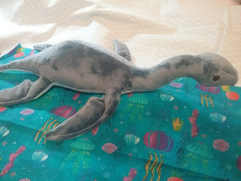 May include: A gray plush toy shaped like a plesiosaur, a long-necked marine reptile, lying on a blue fabric with a sea-life pattern. The fabric has a variety of sea creatures, including jellyfish, seashells, and seaweed.