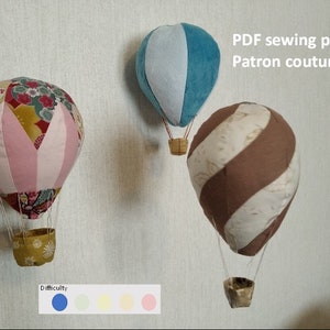 May include: Three fabric hot air balloons in different colours and patterns. The balloons are hanging from the ceiling. The text "PDF sewing pattern Patron couture PDF" is written on the wall. The text "Difficulty" is written below the balloons with a colour scale.