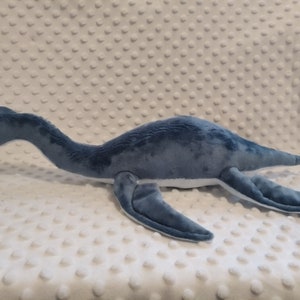 May include: A blue and white plush toy shaped like a plesiosaur, a long-necked marine reptile. The toy has a long neck, a small head with a black eye, and four flippers.