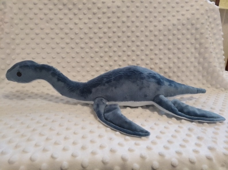 May include: A blue and white plush toy shaped like a plesiosaur, a long-necked marine reptile. The toy has a long neck, a small head with a black eye, and four flippers.