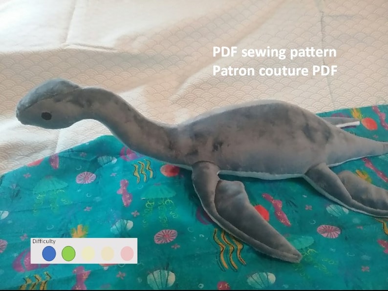 Peut inclure: Un jouet en peluche gris en forme de plésiosaure, un reptile marin préhistorique au long cou et aux nageoires. Le jouet est posé sur un tissu bleu à motif marin. Le texte "PDF sewing pattern Patron couture PDF" est visible dans le coin supérieur droit de l'image. Une échelle de difficulté est représentée sous le jouet avec quatre cercles de couleur : bleu, vert, jaune et rose.