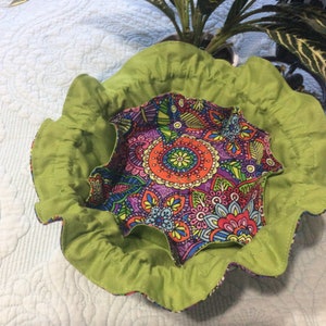 May include: A green fabric bowl with a ruffled edge and a colorful floral print lining. The bowl is perfect for holding small items like jewelry or trinkets.