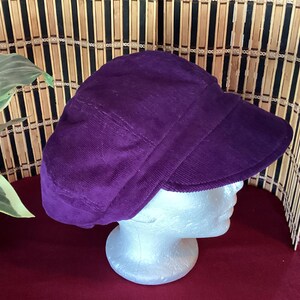 May include: A deep purple corduroy cap with a structured brim. The hat is displayed on a white mannequin head against a backdrop of bamboo and a burgundy surface. The cap's fabric has a ribbed texture.
