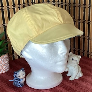 May include: A pale yellow cap with a short brim, displayed on a white mannequin head. The cap has a slightly shiny appearance. Two small ceramic cat figurines are in the foreground, one gray and blue, the other white.