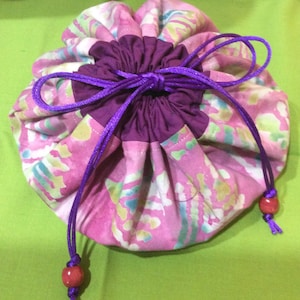 May include: A pink and green drawstring bag with purple drawstrings and red wooden beads. The bag is made of fabric with a batik pattern.