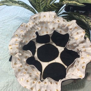May include: A white fabric bowl with a black and gold geometric pattern inside. The bowl has a ruffled edge and gold polka dots.