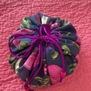 May include: A drawstring bag made of dark blue fabric with a floral print in shades of pink and green. The bag has a purple lining and a drawstring closure with pink and red beads.