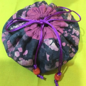 May include: A round drawstring pouch with a floral pattern in shades of purple and navy blue. The pouch is cinched with a bright purple cord, and the ends of the cord are adorned with small, orange beads. The pouch is likely used for storing small items.