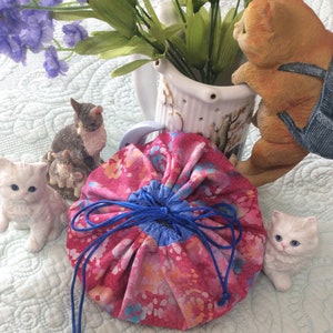 May include: A pink and blue drawstring bag with a floral pattern. The bag is made of fabric and has a blue drawstring. The bag is sitting on a white surface with several small figurines of cats.