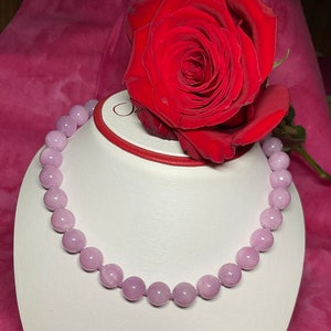 May include: A light purple gemstone necklace with round beads on a white display stand. A red rose is in the background.