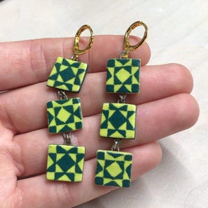 May include: A pair of dangle earrings with three yellow and green geometric patterned squares on each earring. The earrings have gold hoops.