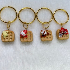 May include: Four gold keychains with different toppings on a waffle charm. The toppings include whipped cream, strawberries, and chocolate syrup.