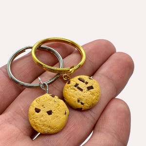 May include: Two gold and silver keychains with chocolate chip cookie charms. The cookies are brown with chocolate chips.