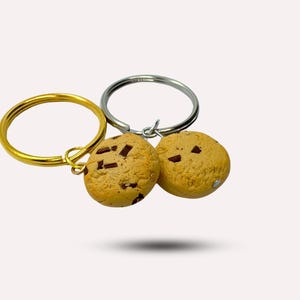 May include: Two chocolate chip cookie keychains, one on a gold key ring and the other on a silver key ring. The cookies are brown and have chocolate chips.